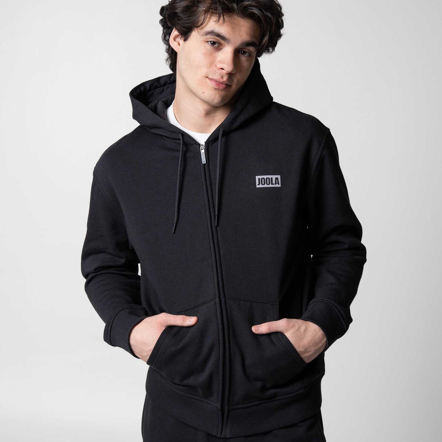 Gray background, image of a male model wearing a zipped up JOOLA Fleece hoodie , black.