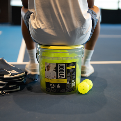 Person sitting on a bucket of JOOLA HC-40 pickleballs.