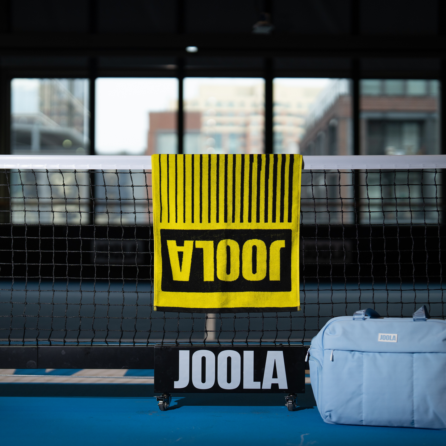 Image of the black and yellow JOOLA wordmark towel handing over a JOOLA Pickleball net at an indoor facility.